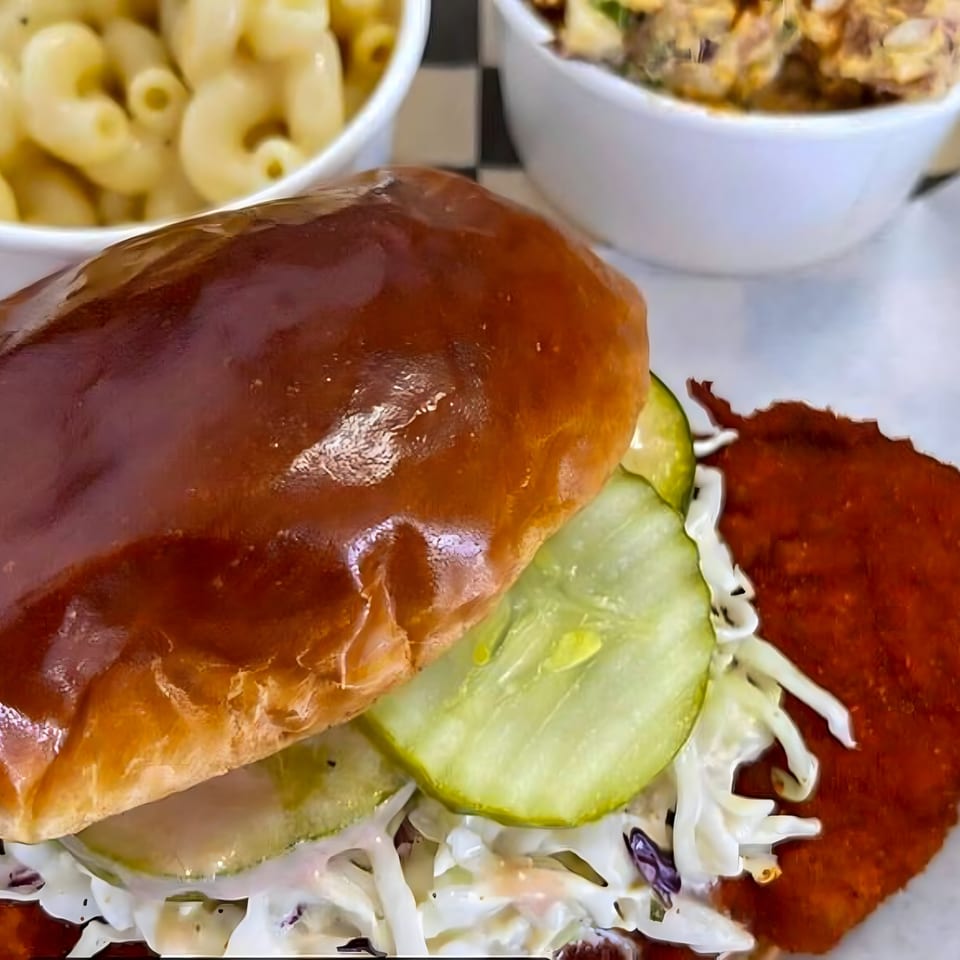 Close-up of a Flew the Coop Nashville Hot Chicken sandwich with pickles, slaw, and sides.