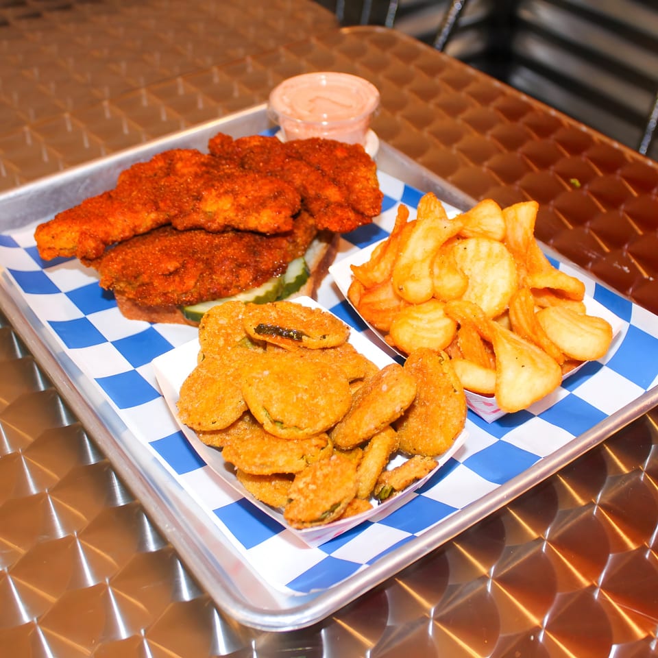 A Nashville hot chicken sandwich, waffle fries, and fried pickles on a metal tray.