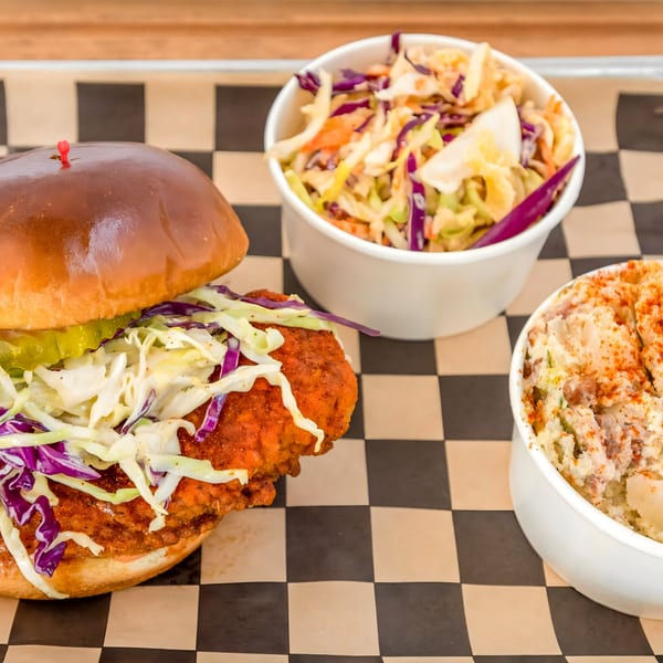 Flew the Coop Nashville Hot Chicken sandwich with coleslaw, pickles, and two sides on checkered paper.