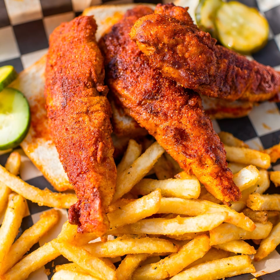 Spicy Nashville hot chicken tenders and crispy fries on a checkered paper liner with pickles.